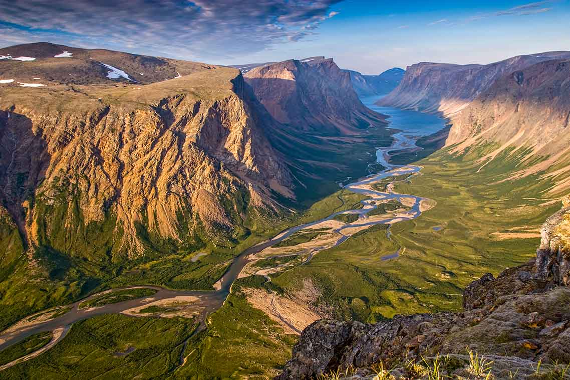 An aerial view of water in a brook weaving and flowing through green vegetation between two tall cliffs into a blue fjord.