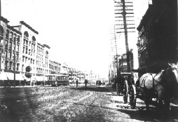 A horse-drawn wagon, streetcar and pedestrians on a street with buildings on either side.