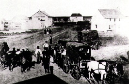 A photo showing people crowded around covered three covered wagons in the street, with five house-like structures in the background.