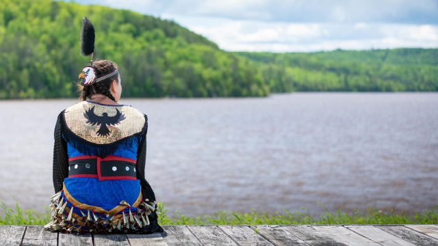 A person in traditional indigenous regalia observing a lake