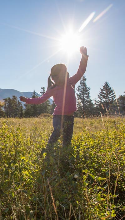 A young Indigenous girl, about five years old, with long hair put up into pigtails dances in a field of wildflowers that comes up to her knees. The background is a sunny blue sky and the sun shows up in a lens flare that crosses the girl's body.