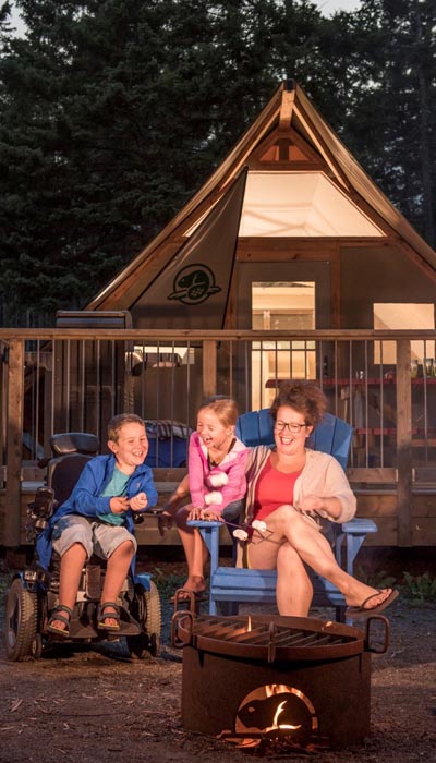 A woman sitting in a blue muskoka chair with two children next to her, laughing. One child is perched on the arm of the muskoka chair. The other is in an electric wheelchair. In front of the group is a metal firepit with a Parks Canada beaver logo cut into the side. Behind them is a semi-permanent tent structure (Otentik) with a wooden frame and a porch. The Otentik has a Parks Canada beaver logo on one flap and is lit inside.  