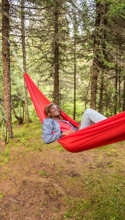 A young Black woman lying in a red hammock attached to a tree in a forest with evergreen trees. She is smiling.
