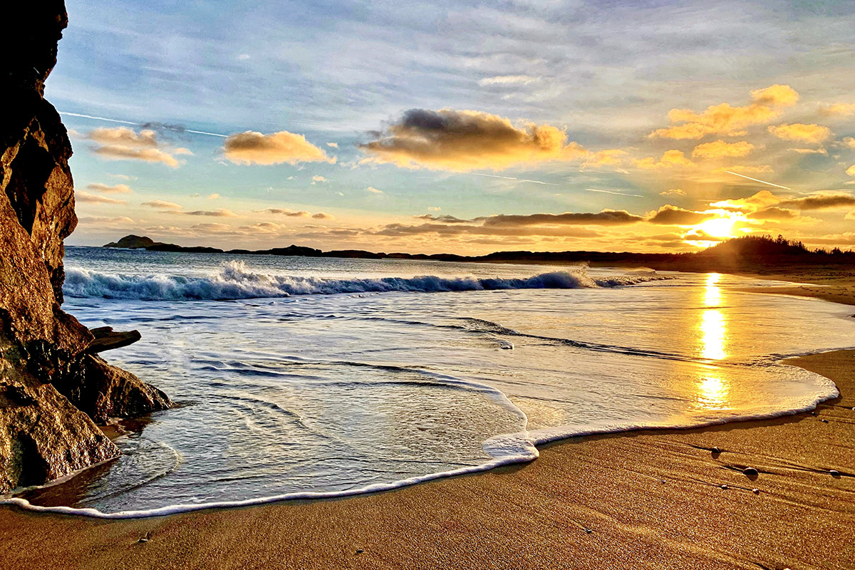 A beach scene at sunset with blues, oranges and yellows in a cloudy sky. The sun is near the horizon casting a glow that reflects off the ocean that extends to the shore. Waves break on the shore, leaving white foam on smooth sand. A rocky formation is visible on the left side of the image. In the distance, the silhouettes of small islands and trees can be seen.