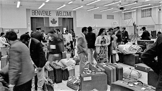 Several people carrying suitcases are gathered in a gymnasium. A Canadian flag and a sign saying, BIENVENUE WELCOME, hang on the back wall.