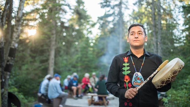 Indigenous man dressed in black with a colourful necklace plays his traditional drum in front of a fire.