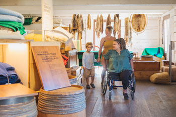 Two adults and a child explore the Storehouse exhibit. One of the adults is using a wheelchair. Behind them, a collection of furs hangs from the exhibit wall.