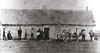 A black-and-white image of 12 people standing in front of a wooden one-storey building. There is a Red River cart in the right side of the image.