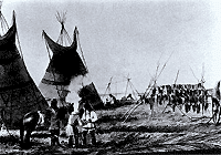 A black-and-white image of a group of people standing with a horse in the foreground, two tipis and a rack made out of wooden sticks with furs draped over it in the background.