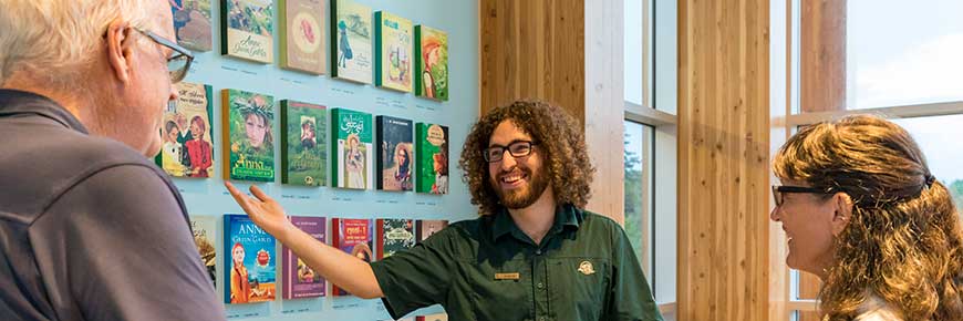 A guide stands in front of the international book cover wall in the Green Gables Visitor Centre Exhibit Hall, talking to a man and woman facing him. 