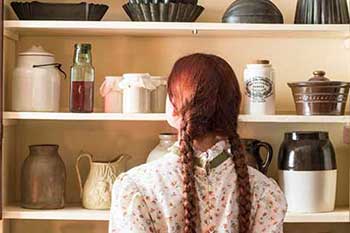 An Anne Shirley impersonator with two red braids stands with her back to the camera in front of an open cupboard filled with cookware at Green Gables Heritage Place. 