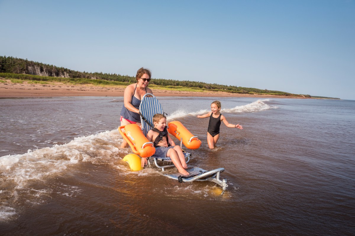 A mother plays with her two children in the waves at the beach. One child is buckled into an assistive device that allows him to go into the water. 