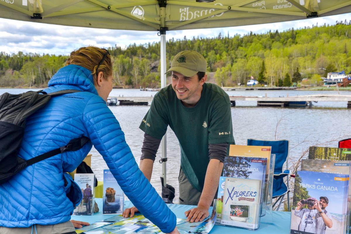 A Parks Canada staff smiles under a booth near the water as they speak with a visitor.