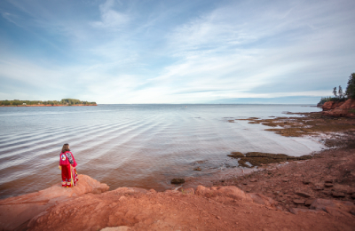 A woman wearing a long red dress, holding a hand drum, stands on shore while looking out into the water.