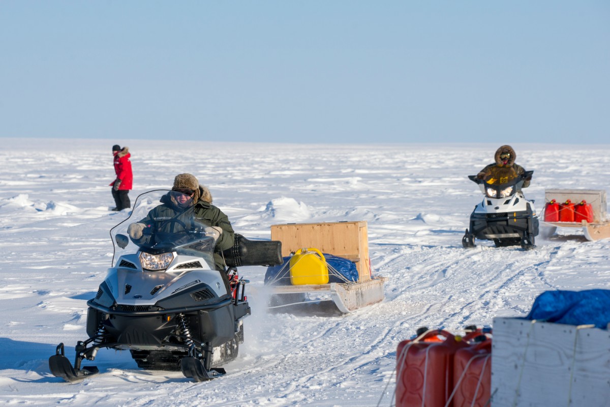Two people ride their snowmobiles while trailing sleds of goods along on a snow-covered tundra.