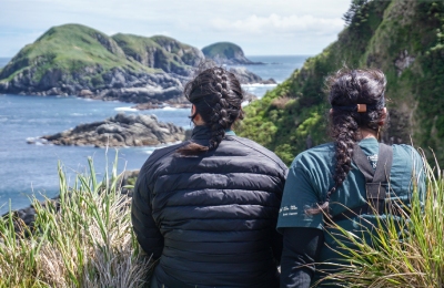 Two women sit on the edge of a cliff that overlooks a coastal vista and islands.