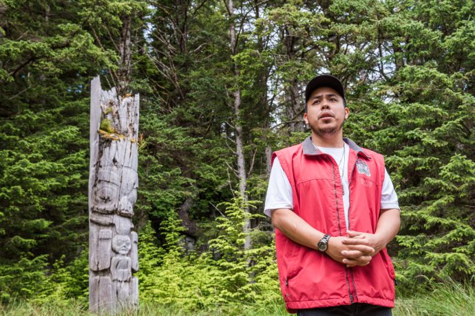 An Indigenous man stands next to the wooden poll as he speaks.
