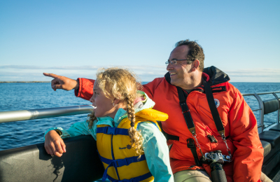 A father points to the water from aboard a boat while his daughter next to him looks excitedly in that direction.