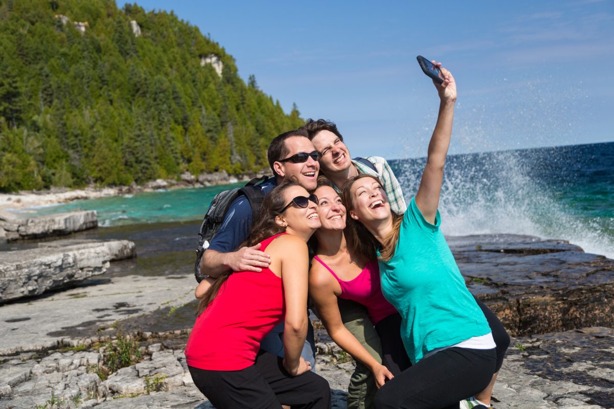 A group of 5 adults gather around one of them who is taking a group selfie near the water’s edge.