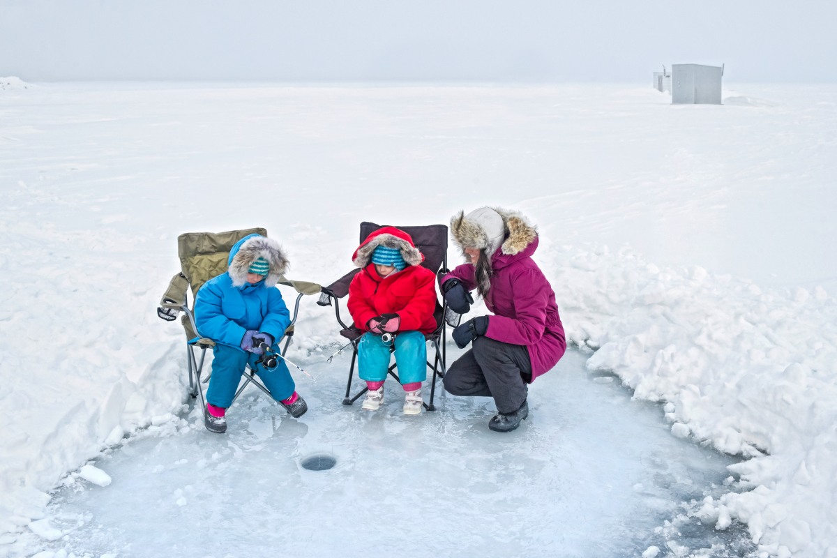 A mother and her two children are bundled up on a flat snow-covered landscape while gathered around an ice fishing hole.