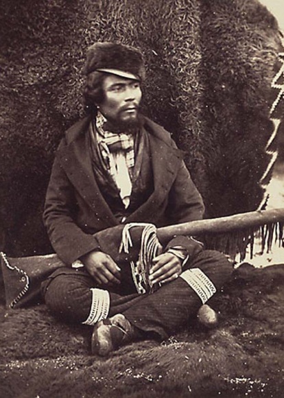 A vintage photo of a man sits cross legged against a pile of bison hide holding his rifle.