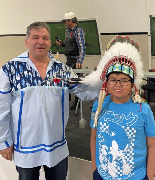 A smiling man with a poppy pinned to a traditional white shirt with blue ribbons and feathers stands next to a boy with glasses wearing a feather and beaded headdress and blue t-shirt.