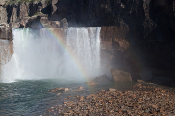 A small waterfall with a rainbow above and a pebbled beach in the foreground.