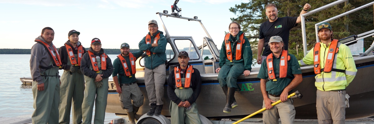A group of 10 Parks Canada staff and Indigenous community members smile on the shore in front of their electrofishing boat that’s being launched on a trailer.