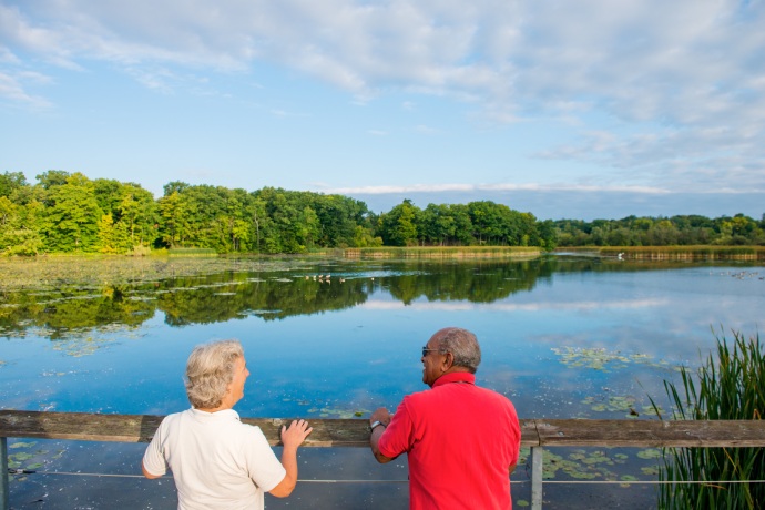 Two senior people stand next to one another smiling as they look over a wooden rail onto a wetland.