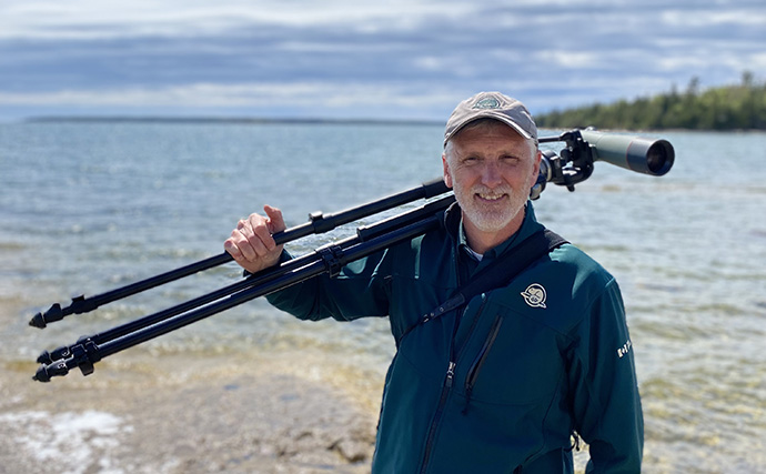 A Parks Canada employee stands and smiles on the shore while carrying a tripod with a telephoto lens attached to it.
