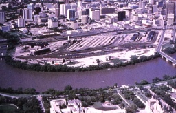 An aerial photo of railyards with a river in the foreground and tall office buildings in the background.