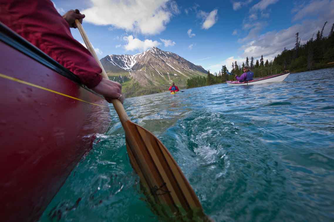 A person paddling a red canoe with two kayakers ahead, forest on the shore to the right and a mountain in the distance ahead. 