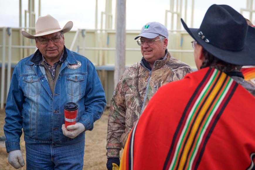 A man wearing a jean jacket and a cowboy hat speaking with a man wearing a ceremonial blanket and another man in a Wildlife Conservation Society baseball cap.