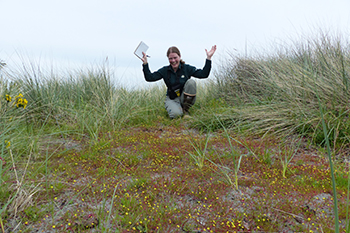 Park biologist raises her arms in celebration near small yellow flowers growing in the sand.