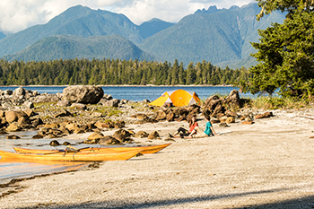 A man and a woman sitting on a beach next to a yellow tent and two kayaks