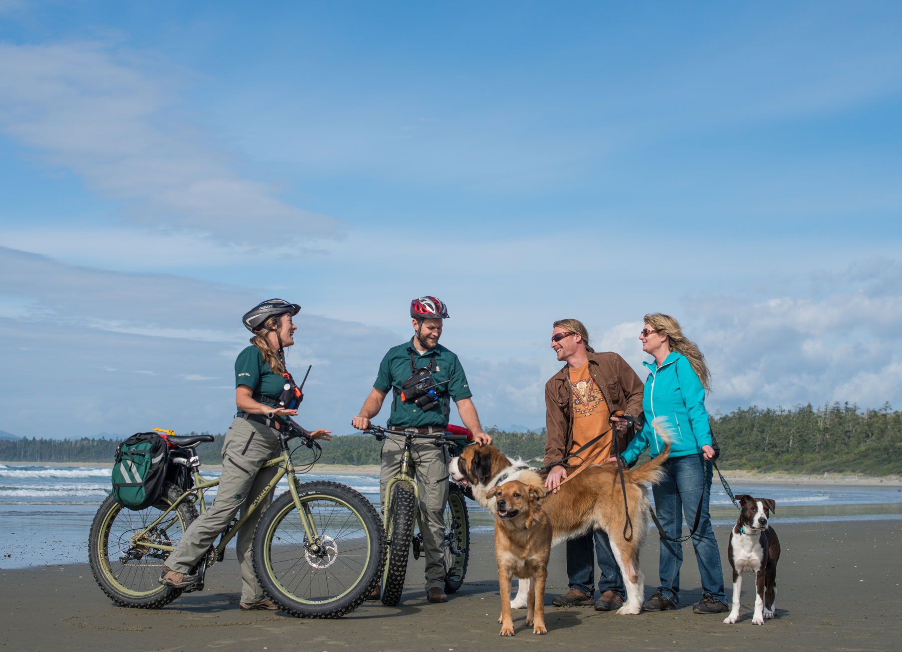 Parks Canada staff on bicycles talking with a couple on the beach with three dogs on leash
