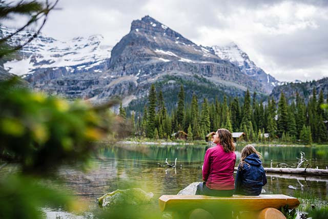 A woman and child sit on a log bench on the shore of Lake O’Hara enjoying the mountain views