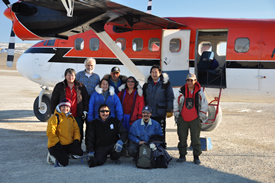 A group of people pose on a landing strip by the open door of a twin otter aircraft