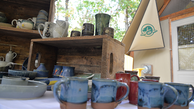 The art of Trish MacDonald, local cermaics artist, neatly displayed in front of a Parks Canada roofed accommodation; the oTENTik. The display features colourful pottery mugs and serving ware.