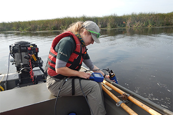 A member of the resource conservation team uses a piece of specialised equipment to collect data on the water quality in the marsh. The employee holds the equipment out of a motor boat and into the water with one hand, while reading data off a screen held in the other. 