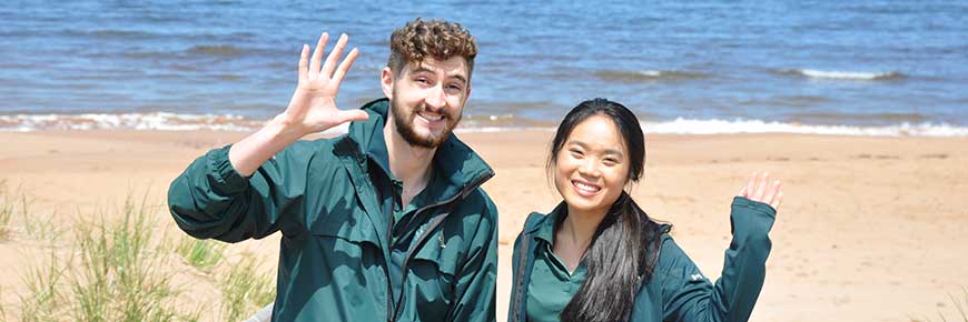 Two Parks Canada students in uniform stand and wave at the entrance to Dalvay Beach, PEI National Park. 