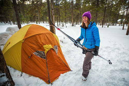 A woman holding skis smiles in front of her tent