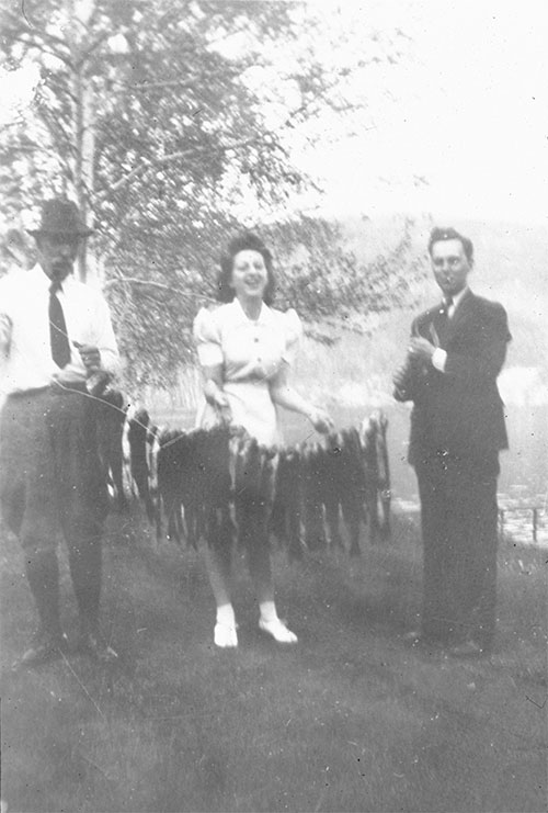 A woman, accompanied by two men, holds several dozen fish in her hands at lac à la Pêche around 1940