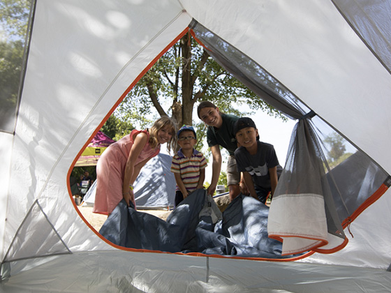 Three children and a Parks Canada employee smiling and looking inside a tent at a Learn-to Camp outreach event. The children are helping the employee set up a tent.