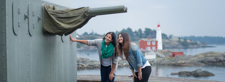 Two young women observe Belmont Batterie’s twin-barrelled 6-pounder gun with Fisgard Lighthouse in the background.