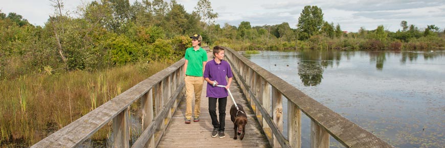 Two visitors and a dog walk along a boardwalk in a national park. The dog is on a leash.
