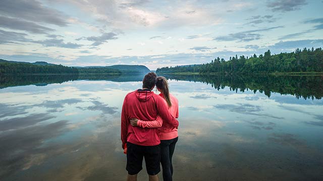 Two adults holding each other by the waist look out over a calm, forested lake in La Mauricie National Park.