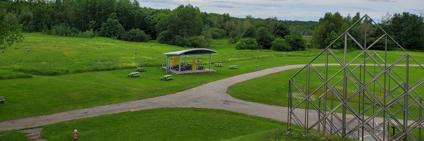A view of the picnic shelter, surrounding picnic benches and field of milkweed among a green lawn on a cloudy day. 
