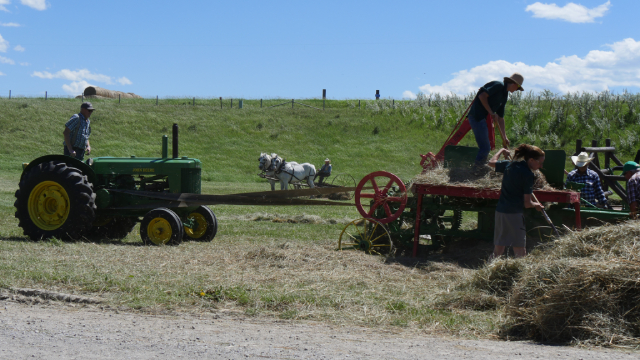 People on tractors and farm equipment with a grassy field and blue skies in the background. 