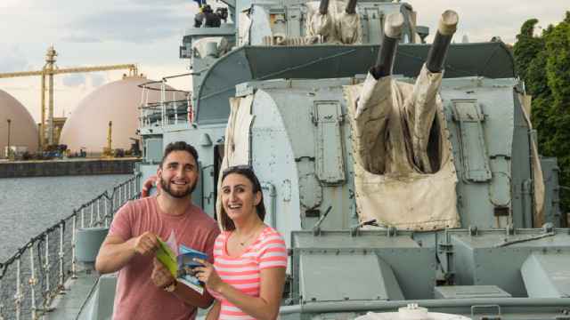 Visitors hold a vistor guide and smile at the camera on board the HCMS Haida destoyer. 